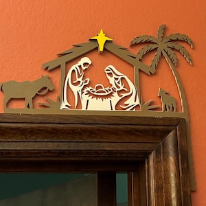 以下が含まれることがあります： 両側にヤシの木のある金色の金属製のクリスマスの誕生シーン。シーンは、飼い葉桶にいるマリア、ヨセフ、そして赤ちゃんのイエスを描いています。シーンは茶色の木製の表面に取り付けられています。