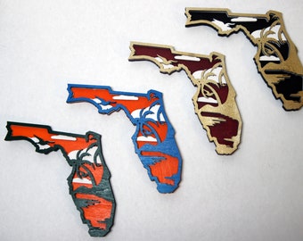 May include: Four wooden Florida state outline ornaments, each painted in a different color: black, maroon, blue, and green. Each ornament features a white silhouette of palm trees and a sunset.