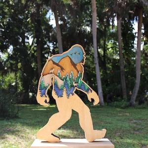 May include: A wooden cutout of Bigfoot walking through a forest scene. The cutout is painted with a blue sky, brown mountains, green trees, and a deer.