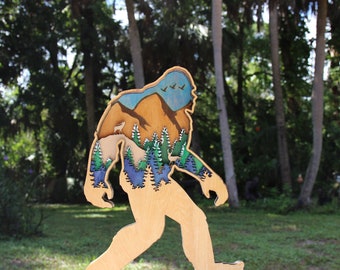 May include: A wooden cutout of Bigfoot walking through a forest scene. The cutout is painted with a blue sky, brown mountains, green trees, and a deer.