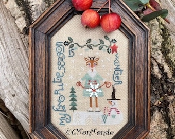 Puede incluir: Un diseño de punto de cruz enmarcado que presenta un ciervo con un vestido azul sosteniendo un copo de nieve, con un muñeco de nieve y un árbol en el fondo. El texto "My favorite time of season" está bordado en el lado izquierdo del diseño, y "Winter" está bordado en el lado derecho. La parte inferior del diseño está firmada "C Mon Monde".