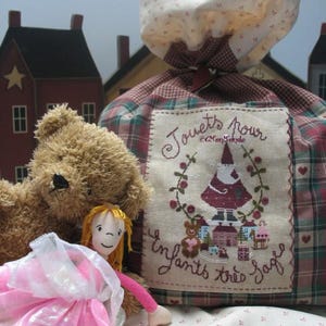 Peut inclure: Un grand sac à jouets en tissu à carreaux avec une fermeture à cordon, avec un patch cousu avec les mots "Jouets pour Enfants très sage" et une image décorative. Un ours en peluche et une poupée sont au premier plan.