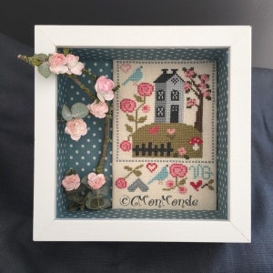 May include: A framed cross-stitch design featuring a small house with a green roof, a fence, and a tree with pink blossoms. The house is gray with white trim and a black door. The design is surrounded by a blue and white polka dot border. The text "C Mon Monde" is visible at the bottom of the design.