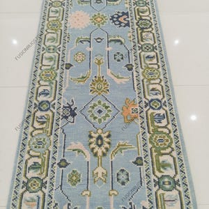 May include: A light blue runner rug with an intricate floral and geometric pattern. The rug features a central design with various shapes in shades of green, white, and peach. The border is a repeating pattern of squares and floral motifs.