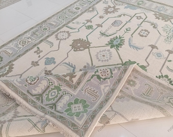 May include: A cream-colored area rug with a detailed floral and geometric pattern in shades of green, blue, and brown. The rug has a border with a repeating design and a folded corner, showcasing the intricate weave.