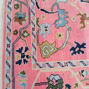 May include: A close-up of a pink rug with a detailed floral pattern. The rug features a border with light blue and navy blue accents. The design includes various floral and geometric shapes in shades of blue, green, and tan.