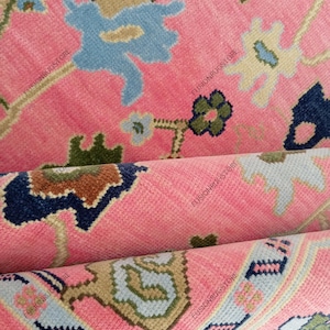 May include: Close-up of a pink rug with a floral pattern. The rug features a repeating design of blue, beige, and green floral motifs on a pink background. The rug is folded, revealing the texture and layers of the fabric.