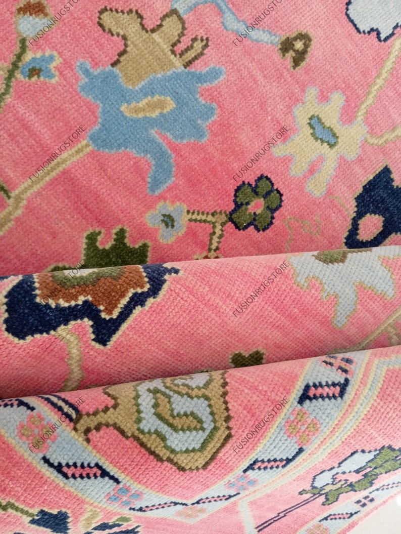 May include: Close-up of a pink rug with a floral pattern. The rug features a repeating design of blue, beige, and green floral motifs on a pink background. The rug is folded, revealing the texture and layers of the fabric.