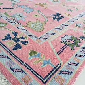 May include: A close-up of a pink rug with a detailed floral pattern. The rug features a central field of pink with blue, green, and brown floral motifs. The border is a darker pink with a geometric design and a light blue accent. The rug has a fringe.