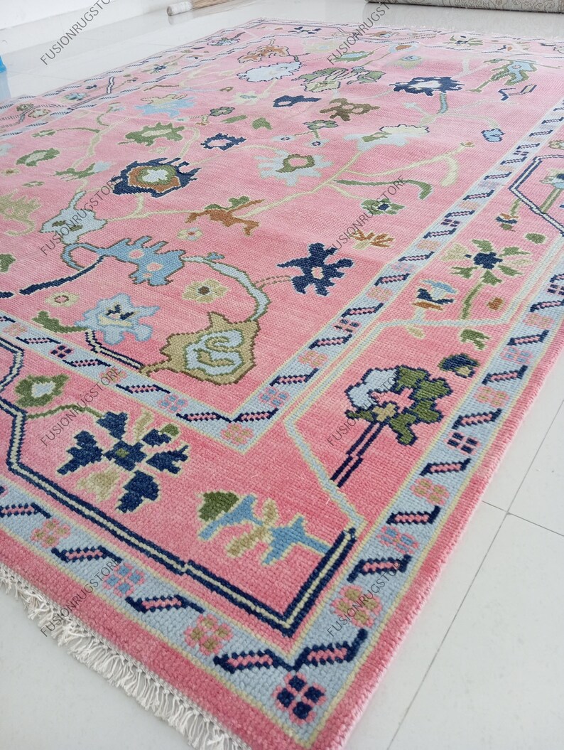 May include: A pink area rug with a floral pattern. The rug features a central field of pink with blue, green, and brown floral motifs. A border of light blue and navy blue frames the rug, with a fringe along one edge.