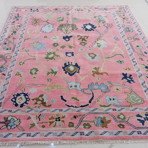 May include: A large, rectangular pink rug with an intricate floral pattern. The rug features a central field of pink with a repeating design of flowers in shades of blue, green, and brown. A detailed border frames the rug.