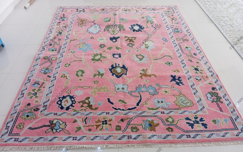 May include: A large, rectangular pink rug with an intricate floral pattern. The rug features a central field of pink with a repeating design of flowers in shades of blue, green, and brown. A detailed border frames the rug.