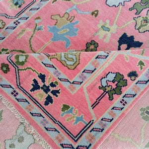 May include: A pink rug with a floral pattern and a geometric border. The rug features light blue, navy, and green accents. The rug is folded over, revealing the underside with a similar design.