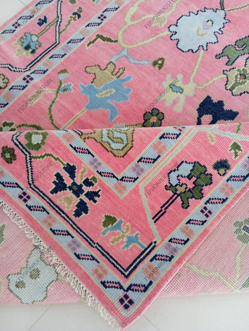 May include: A pink rug with a floral pattern and a geometric border. The rug features light blue, navy, and green accents. The rug is folded over, revealing the underside with a similar design.