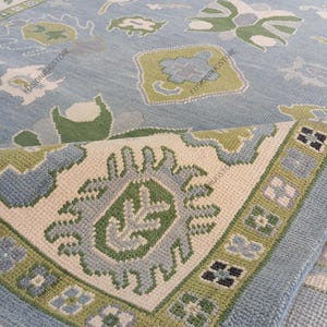 May include: A light blue area rug with a detailed pattern. The rug features a central design with green and gray floral motifs, surrounded by a border of green, blue, and cream colors. The rug is partially rolled, revealing the underside.