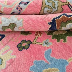 May include: Close-up of a rolled-up pink rug with a floral pattern. The rug features a repeating design with blue, green, and beige accents. The rug's texture is visible, showcasing its woven construction.
