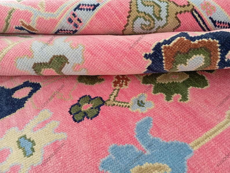 May include: Close-up of a rolled-up pink rug with a floral pattern. The rug features a repeating design with blue, green, and beige accents. The rug's texture is visible, showcasing its woven construction.