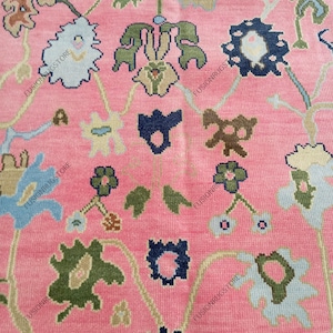 May include: Close-up of a pink rug with an intricate floral pattern. The design features stylized flowers in shades of blue, green, and brown, connected by curving vines. The rug's background is a solid, soft pink, creating a vibrant contrast with the detailed floral elements.