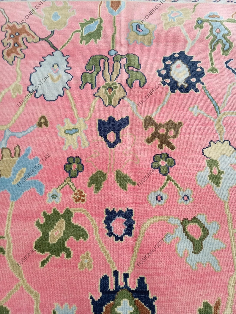May include: Close-up of a pink rug with an intricate floral pattern. The design features stylized flowers in shades of blue, green, and brown, connected by curving vines. The rug's background is a solid, soft pink, creating a vibrant contrast with the detailed floral elements.