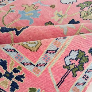 May include: Close-up of a pink rug with a floral pattern. The rug features a repeating design of flowers and geometric shapes in shades of blue, green, and beige. The rug is folded, revealing its layered construction.