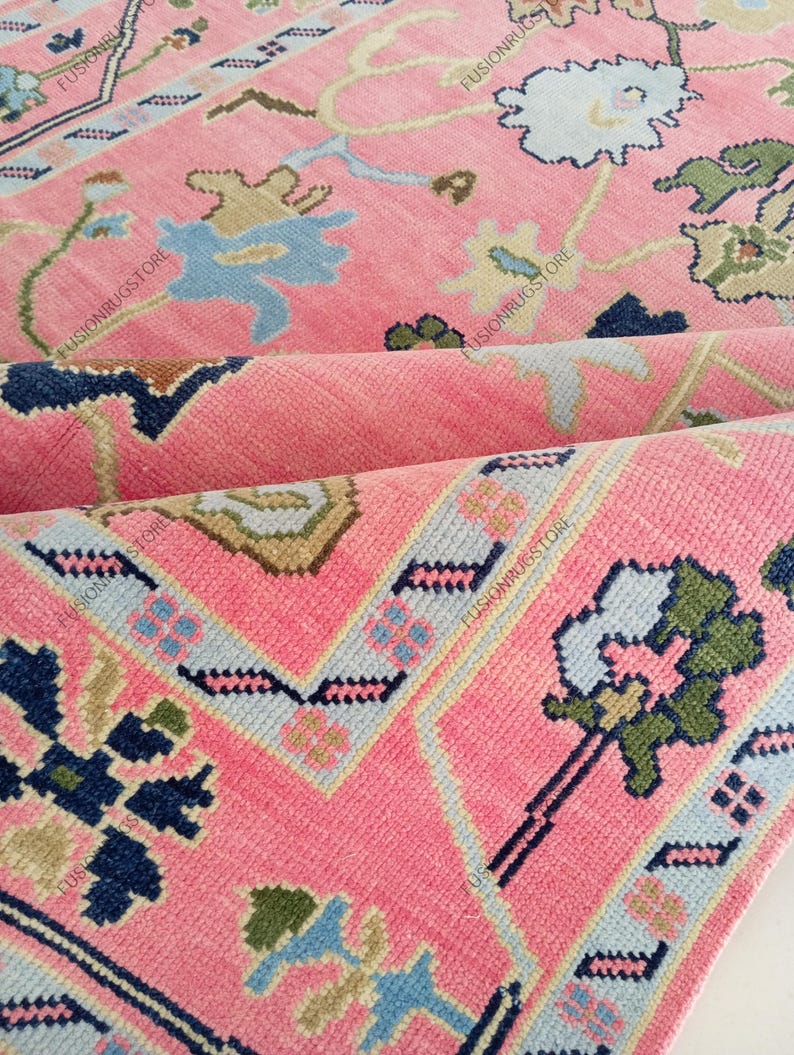 May include: Close-up of a pink rug with a floral pattern. The rug features a repeating design of flowers and geometric shapes in shades of blue, green, and beige. The rug is folded, revealing its layered construction.