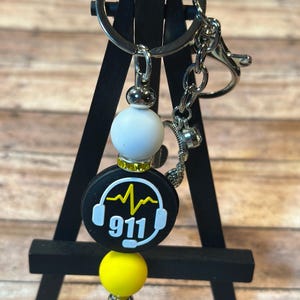 May include: A keychain with a silver ring and clasp. The keychain features a black charm with a white 911 graphic and yellow heartbeat design, white and yellow beads, and a silver headphone charm.