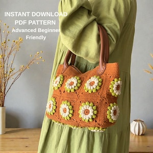 May include: An orange crochet tote bag with white and green flower appliqués. The bag has brown leather handles and the text "INSTANT DOWNLOAD PDF PATTERN Advanced Beginner Friendly".