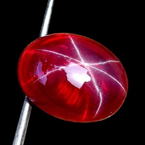 Pode incluir: Uma gema de rubi estrela vermelho de formato oval com um padrão de estrela de seis pontas distinto em sua superfície de cabochão lisa e polida. A pedra carmesim é segurada por uma ferramenta de metal prateado contra um fundo preto.