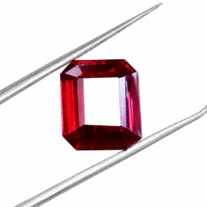 Puede incluir: Una gema de color rojo intenso y vibrante con un corte esmeralda rectangular, que presenta superficies facetadas que reflejan la luz. La gema se sostiene de forma segura entre las dos puntas metálicas plateadas de unas pinzas.