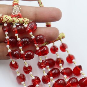 May include: A multi-strand necklace featuring translucent red teardrop-shaped beads interspersed with small white spacer beads. The strands are gathered at the top by a gold-coloured braided cord connector with red thread accents.
