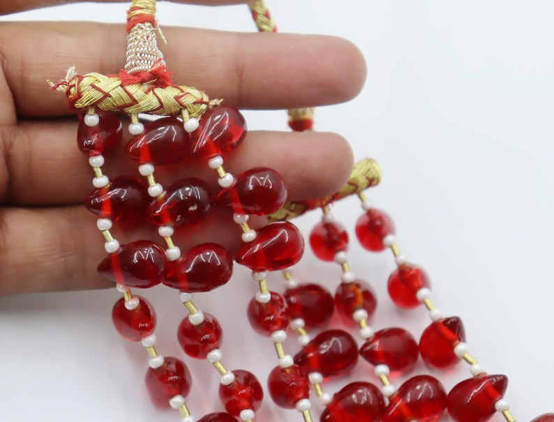 May include: A multi-strand necklace featuring translucent red teardrop-shaped beads interspersed with small white spacer beads. The strands are gathered at the top by a gold-coloured braided cord connector with red thread accents.