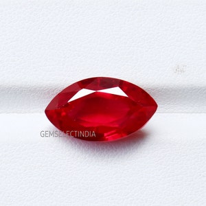 Può includere: Una gemma sfaccettata a taglio marquise in una vibrante tonalità rossa. La gemma è posta su uno sfondo bianco. Il testo "GEMSELECTINDIA" è visibile in basso a sinistra.