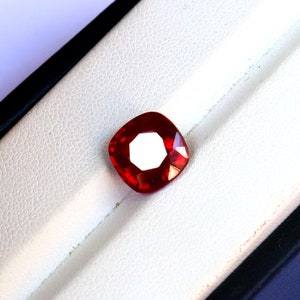 May include: A faceted, cushion-cut, red gemstone. The gem is set against a white, textured background within a black-trimmed box. The gem's facets reflect light, creating a bright, central highlight.