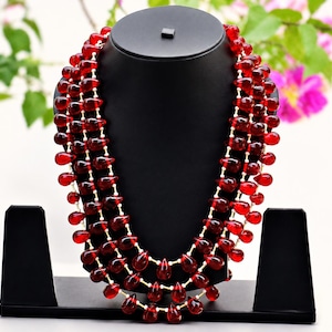 May include: A multi-strand necklace featuring numerous deep red, teardrop-shaped beads strung on thin, light-colored cord. The beads are arranged in three distinct, cascading layers that drape symmetrically around the neck display form.