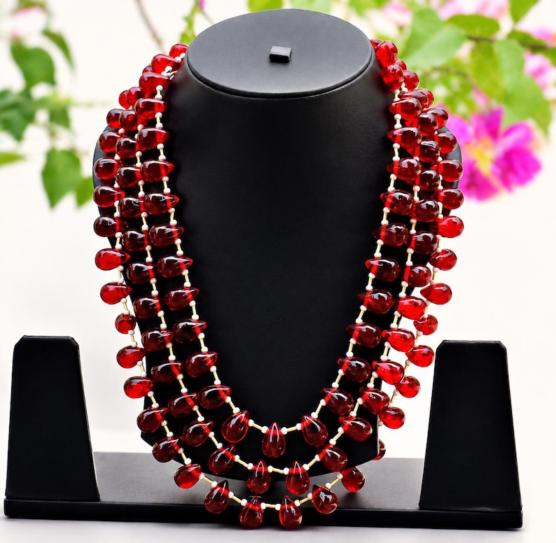May include: A multi-strand necklace featuring numerous deep red, teardrop-shaped beads strung on thin, light-coloured cord. The beads are arranged in three distinct, cascading layers that drape symmetrically around the neck display form.