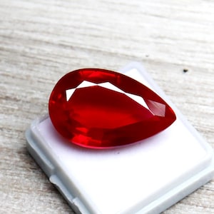May include: A vibrant, pear-shaped, faceted ruby gemstone. The gem is a deep, saturated red color, with a brilliant, reflective surface. It is set on a white plastic base, showcasing its clarity and color. The gemstone is a focal point, suggesting jewelry or collectible.