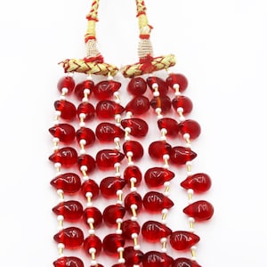 May include: A multi-strand necklace featuring numerous translucent red teardrop-shaped beads interspersed with small white cylindrical beads. The strands are gathered at the top by two gold-toned metallic cylinders with red thread accents.