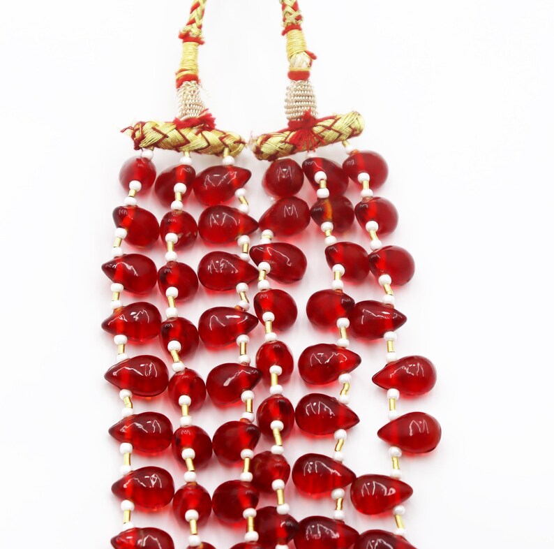 May include: A multi-strand necklace featuring numerous translucent red teardrop-shaped beads interspersed with small white cylindrical beads. The strands are gathered at the top by two gold-toned metallic cylinders with red thread accents.