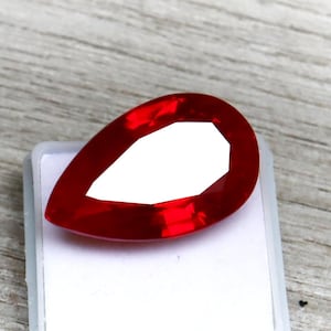May include: A vibrant, pear-shaped, faceted red gemstone. The gem is set against a white background, showcasing its deep red color and cut. The gemstone is likely a ruby, with a polished surface.