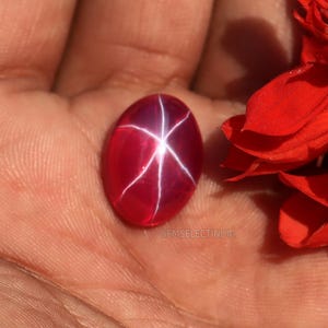 May include: A red star ruby gemstone with a star-like pattern in the center. The gemstone is oval-shaped and has a smooth, polished surface.