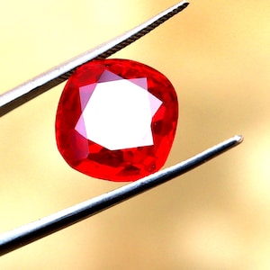 May include: A vibrant red gemstone, held by silver tweezers, against a warm, golden background. The gem has a cushion cut with a large, white, geometric center. The facets reflect light, showcasing its clarity and brilliance.