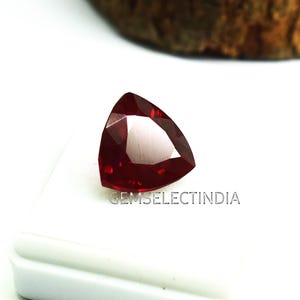 Pode incluir: Uma gema de cor vermelha profunda com um corte triangular, exibida em uma superfície branca. A gema é facetada e tem um alto brilho. O texto "GEMSELECTINDIA" é visível abaixo da gema.