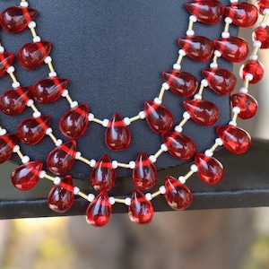 May include: A multi-strand necklace featuring translucent, deep red, teardrop-shaped beads arranged in three cascading layers. The red beads are separated by small, cylindrical white spacers and thin gold-toned metal wire links.