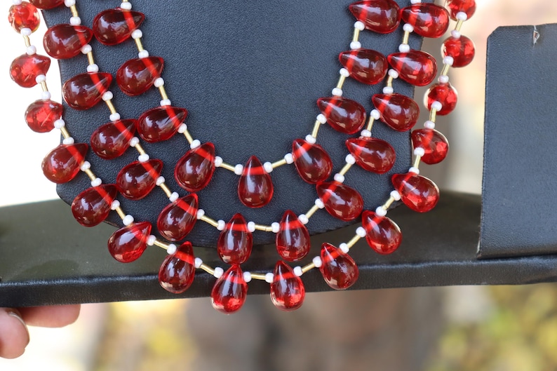 May include: A multi-strand necklace featuring translucent, deep red, teardrop-shaped beads arranged in three cascading layers. The red beads are separated by small, cylindrical white spacers and thin gold-toned metal wire links.