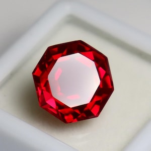 Puede incluir: Una piedra preciosa de color rojo vibrante con un corte octogonal, que presenta múltiples facetas triangulares y trapezoidales que crean un patrón simétrico. La piedra muestra un tono carmesí profundo con reflejos brillantes.