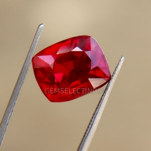 May include: A vibrant red gemstone, held by silver tweezers. The gem has a cushion cut with multiple facets, reflecting light. The image includes the text "GEMSELECTINDIA".