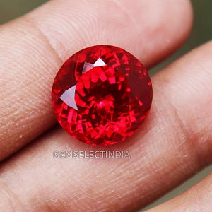 May include: A vibrant red gemstone with a faceted cut. The gemstone is round and has a high polish. The gemstone is held in a hand.