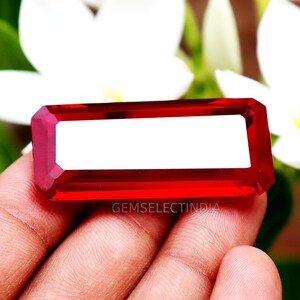 Könnte beinhalten: Ein rechteckiger, facettierter, roter Edelstein mit weißer Mitte. Der Edelstein wird in einer Hand gehalten, mit dem Text "GEMSELECTINDIA" sichtbar. Der Hintergrund zeigt weiße Blumen und grünes Laub.