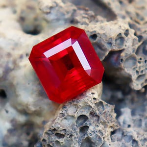 Può includere: Una gemma rossa vibrante, tagliata a forma quadrata con sfaccettature. La gemma è posizionata su una superficie rocciosa grigia e strutturata. Le sfaccettature della gemma riflettono la luce, creando riflessi bianchi brillanti. La composizione generale mette in risalto il colore e la chiarezza della gemma.