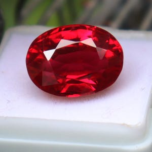 May include: A vibrant, oval-cut gemstone with a deep, saturated red color and multiple facets that reflect light. The stone displays high clarity and a polished surface, showcasing a brilliant luster throughout its entire structure.